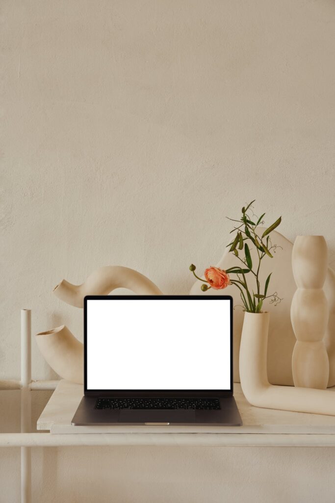 Netbook with white screen placed on shelf near uneven curved ceramic vases with flowers with green leaves near pipes in light room with white wall